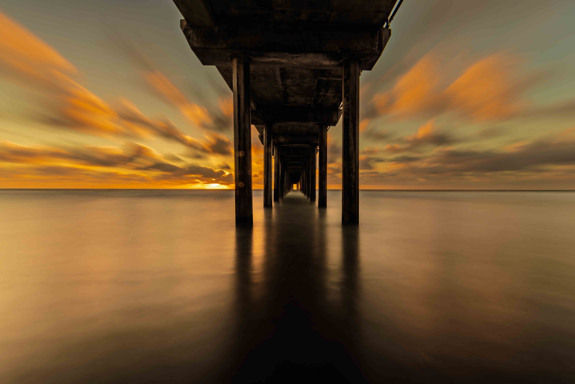Scripps Pier at golden hour, La Jolla — long exposure turns crashing waves to silk beneath weathered wooden pillars against a burning sky