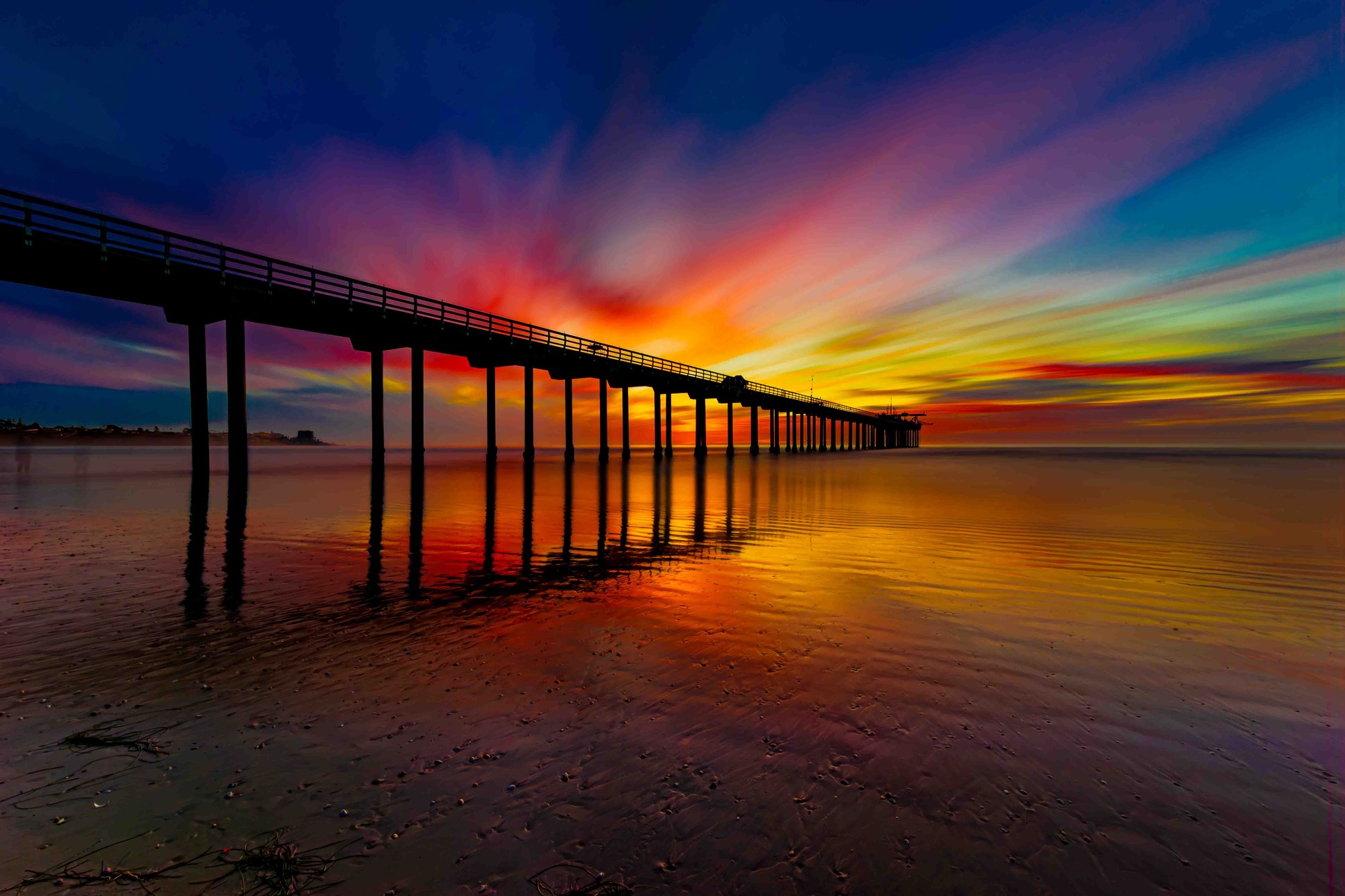 Long-exposure photograph of an iconic San Diego pier at golden hour, waves transformed to silk beneath weathered wooden pillars