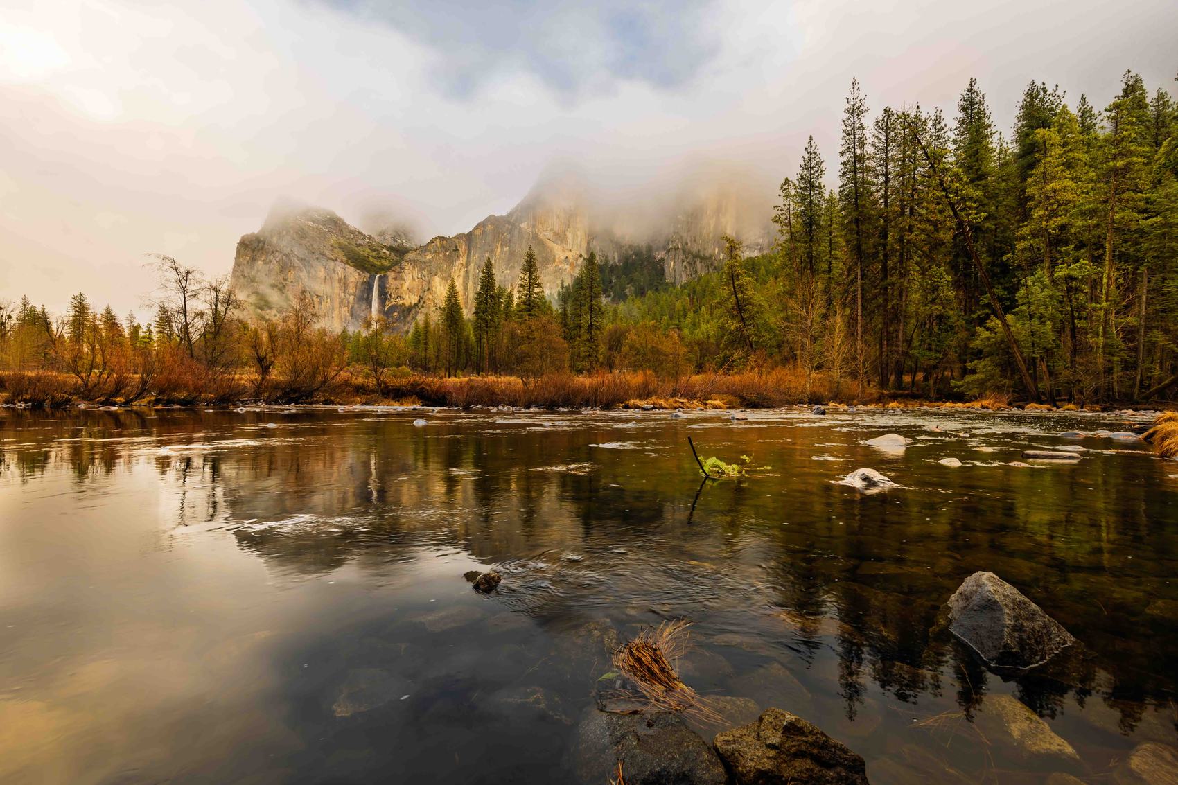 Merced River in Yosemite Valley at dawn — granite walls emerging from morning mist, Bridalveil Fall threading through the clouds, ancient forest reflected in still water
